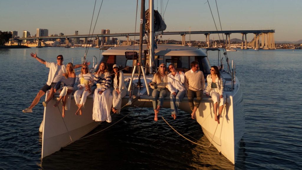 A Group Of People Sits On A Catamaran At Sunset. The City Skyline And Bridge Are In The Background. They Enjoy A Peaceful Moment On The Water.