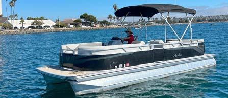 A person steering a pontoon boat on calm blue water with a clear sky and coastline in the background.