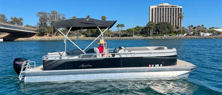 A person steering a pontoon boat on calm waters with a clear sky and a building in the background.