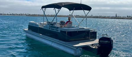 A pontoon boat with a canopy on calm waters under a partly cloudy sky with a person aboard.