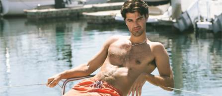 A young man lounges on a boat dock, wearing bright orange swim shorts, with calm waters and yachts in the background.