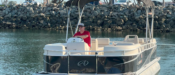A man in a red shirt drives a pontoon boat on calm water, with rocky shoreline and parked vehicles in the background.