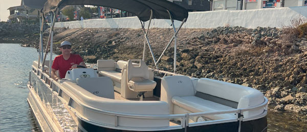 A person in a red shirt steers a pontoon boat on calm waters near a rocky shoreline, under a canopy sunshade.