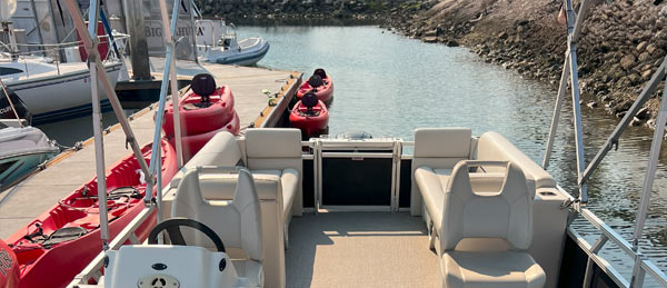 View from the back of a pontoon boat facing a calm marina, with red kayaks docked nearby and other boats moored along the shore.