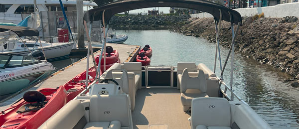 View from the back of a pontoon boat facing a calm marina, with red kayaks docked nearby and other boats moored along the shore.