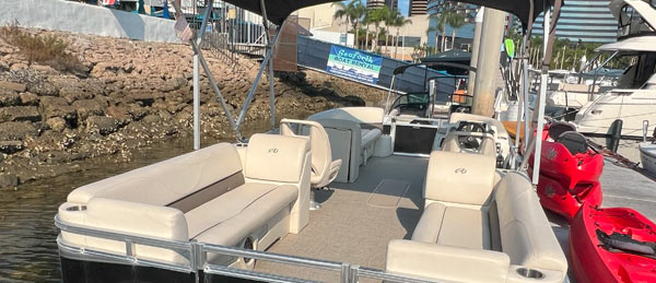 A spacious, beige pontoon boat docked, featuring comfortable seating and a scenic view of palm trees and a waterfront.