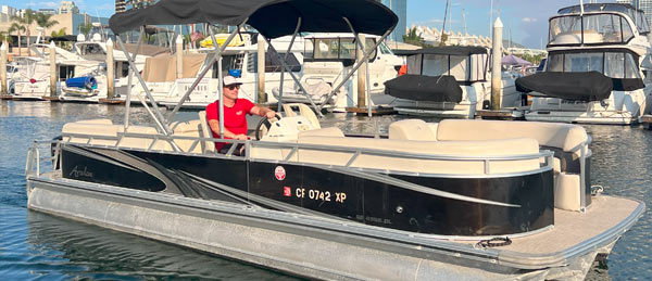 A man in a red shirt and hat steers a black pontoon boat in a marina surrounded by other parked boats.