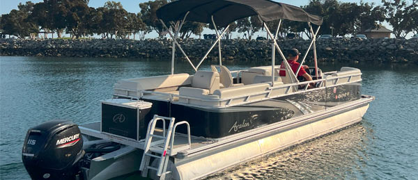A person in a red shirt sits on a luxury pontoon boat with a canopy, moored in calm waters near a rocky shoreline.