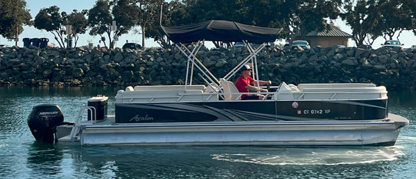 A man in a red shirt navigates a black and silver pontoon boat on calm water, with rocky shore and trees in the background.