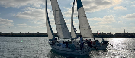 Two sailboats glide through shimmering water under a cloudy sky, with people onboard enjoying a day of sailing.