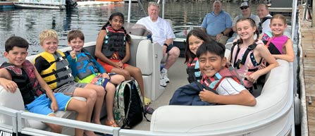 Group of children wearing life jackets sitting on a boat with adults behind them.