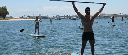 A group of people paddleboarding on a sunny day; a man triumphantly raises his paddle, while a woman balances behind him.