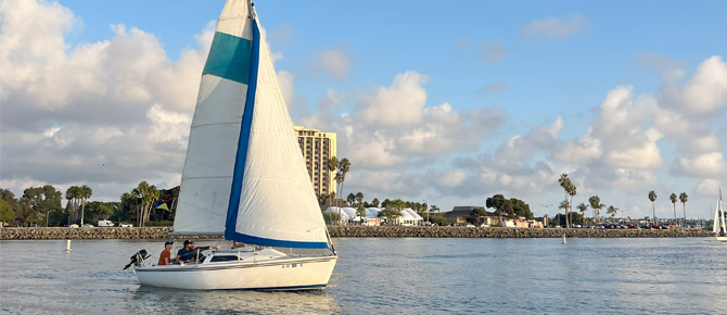 A sailboat with a blue and white sail glides on calm water near a coastline with buildings and trees.