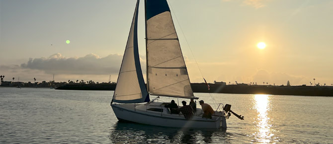 A sailboat with people aboard at sunset, reflecting over calm water.