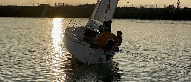 A sailboat with two people on board glides on calm water at sunset, reflecting light on the water.