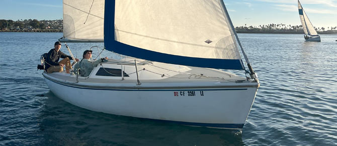 Two people sailing a white sailboat, with another sailboat in the distance on calm waters.