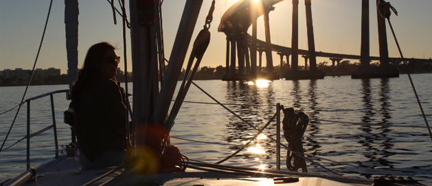 Person on a sailboat at sunset with a bridge silhouette in the background.