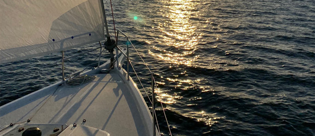 Bow of a sailboat on the water at sunset with sunlight reflecting on waves.