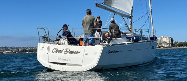 Four people sailing on a yacht named "Cosmic Dancer" off the coast on a clear day.