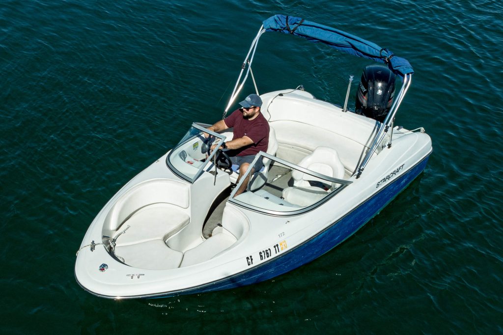 A Man Steers A Small White Motorboat With A Blue Canopy On Clear Blue Water, Surrounded By Sunlit Reflections.