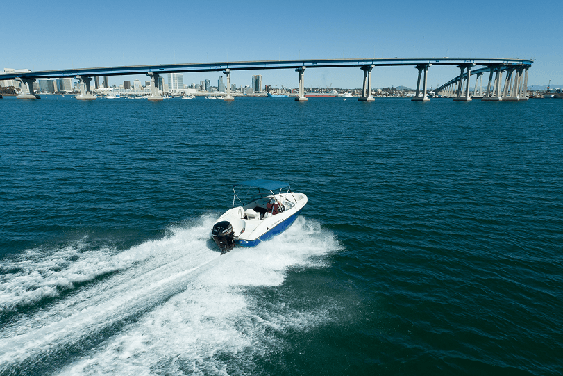 A motorboat speeds through blue waters beneath a bridge, with a city skyline visible in the background on a clear day.