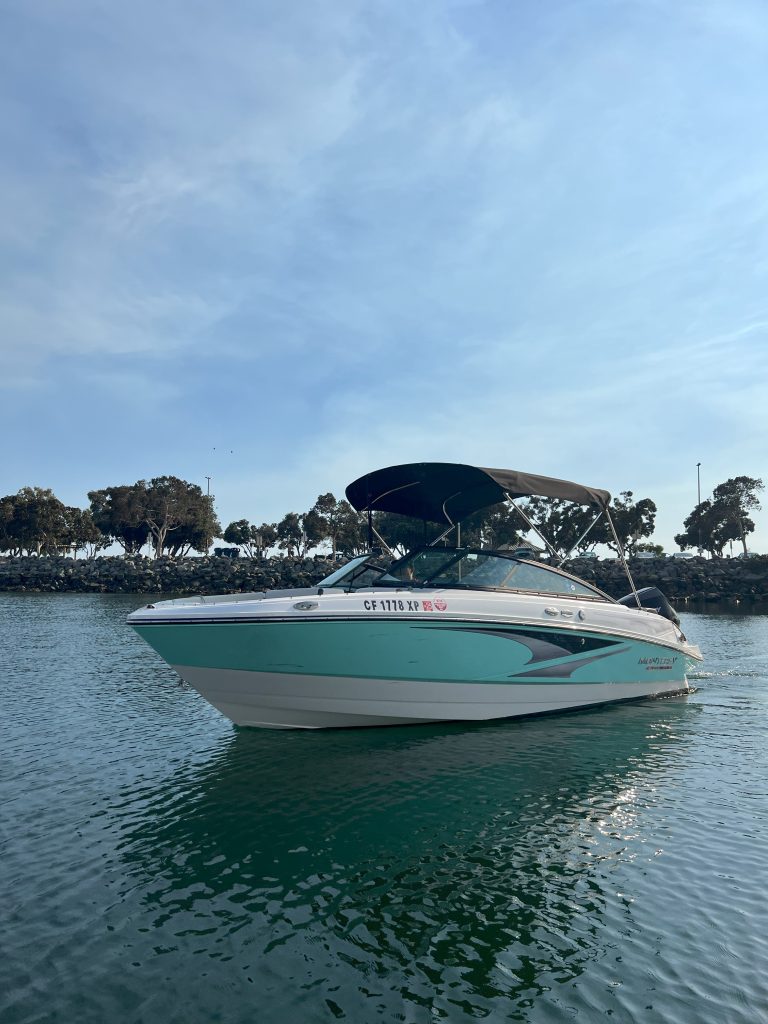 A Man Wearing A Hat Smiles From The Helm Of A Turquoise And White Boat Docked In A Marina, Surrounded By Tall Buildings And Other Boats.
