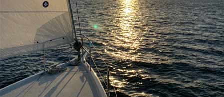 A sailboat glides across the water at sunset, with the sun shining brightly
