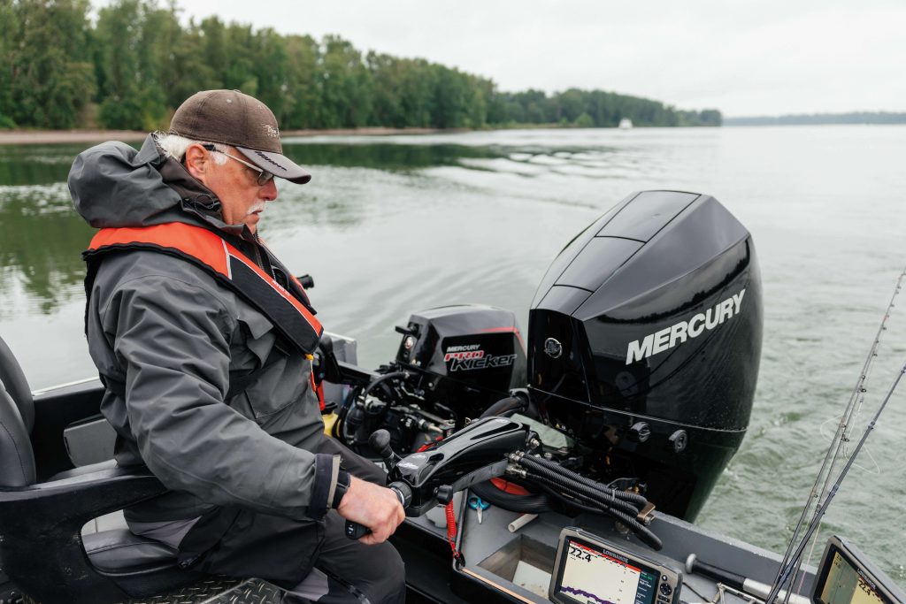 A Person In A Boat Operates A Mercury Outboard Motor, Surrounded By Calm Water And Lush Green Trees Along The Shoreline.