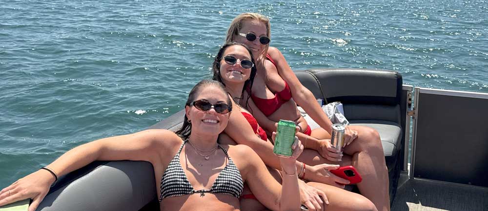 Three women in swimsuits relaxing on a boat with drinks, smiling and enjoying the sunny day on the water