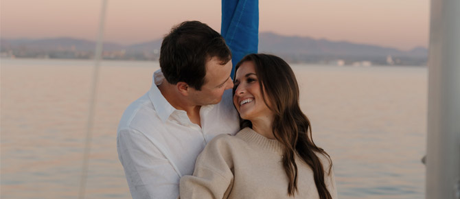A loving couple stands together on a yacht during a sunset bay proposal, capturing the perfect engagement on the water moment.